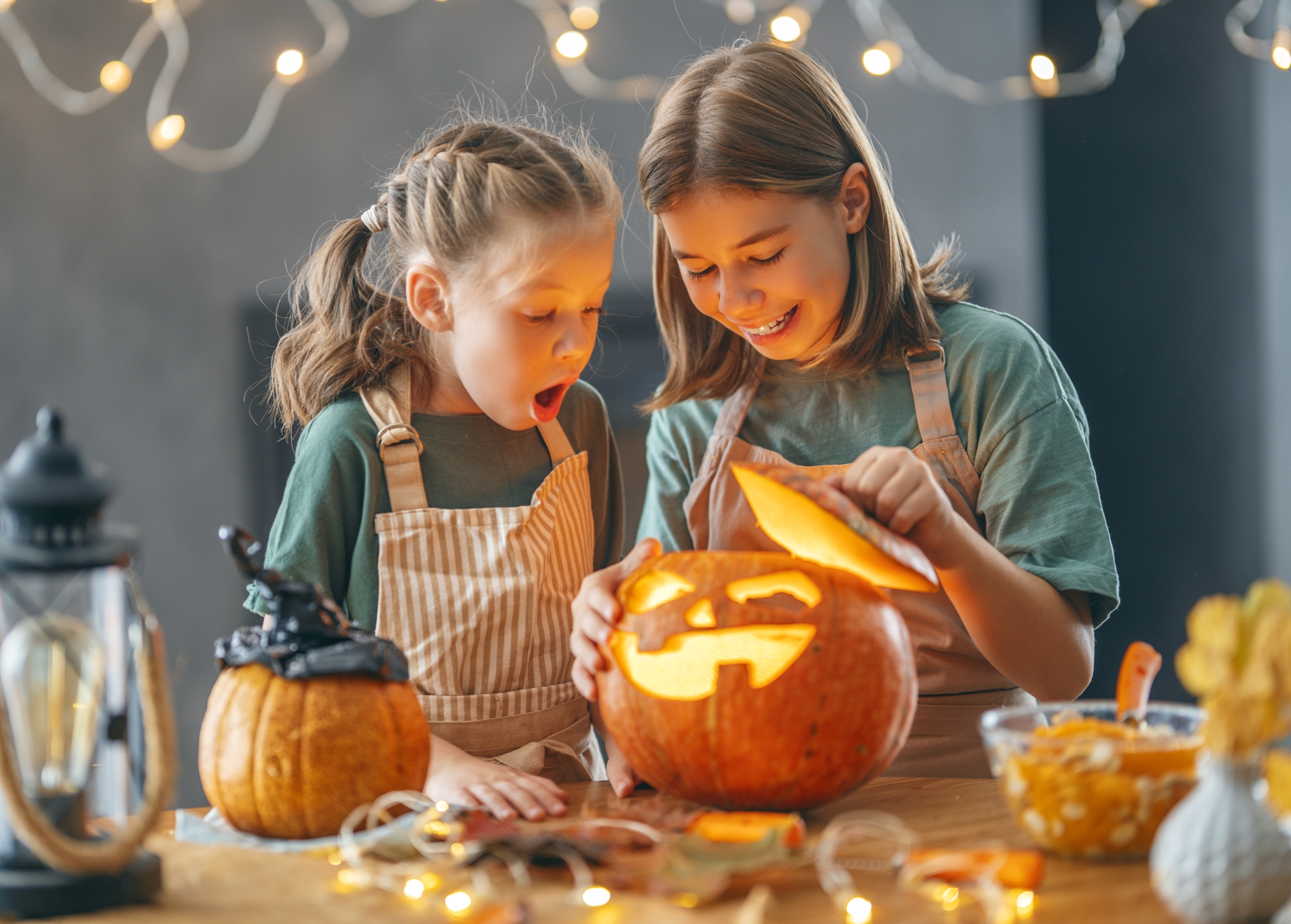 two girls admiring their carved, lit pumpkin - on their kitchen counter wearing tan aprons and soft green shirts - with twinkle lights in foreground and background