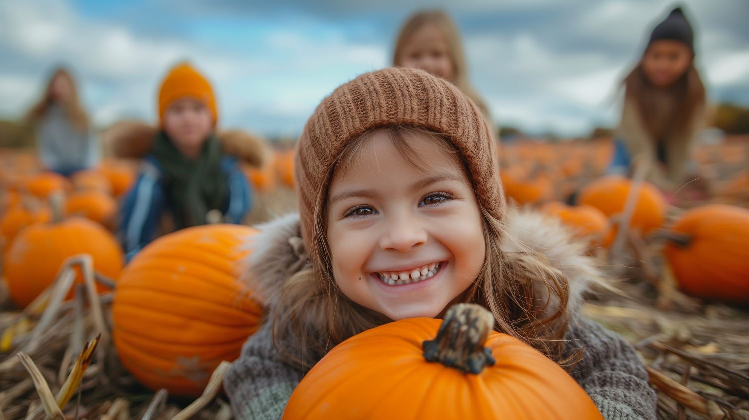 kids in pumpkin field with fall colored knit hats. little girl in foreground laying with chin resting on a pumpkin and smiling - other kids in blurred background surrounded by pumpkins with blue cloudy sky in background