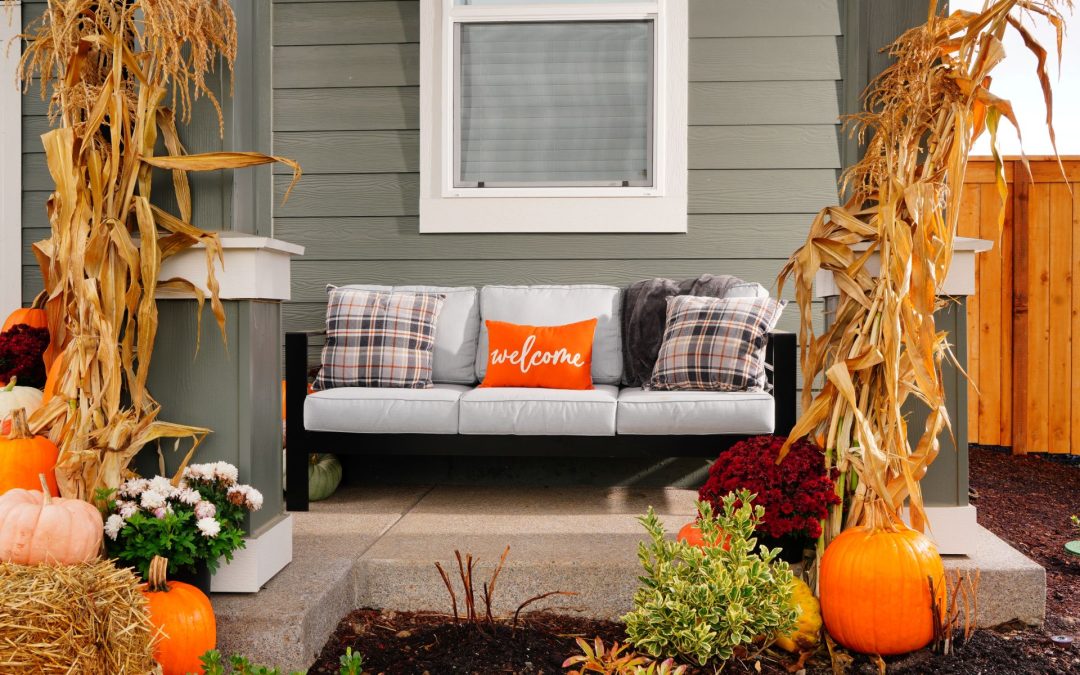 Fall themed porch of new light grey home with white trimmed windows. Two columns painted light and dark grey with white trim. Outdoor couch with very light grey cushions and black iron base. Decor with pumpkins of various sizes and shapes, corn stocks, mums in light pink and burgandy, and hay bales