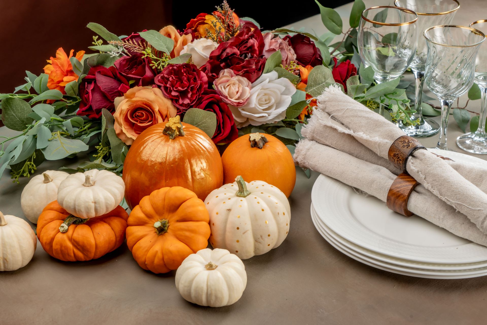 stacked white plates with raw edged linen ivory napkins and wooden holders. centerpiece garland made of eucalyptus and greens base with various roses in fall colors (burgundy, orange, pink, and white). PUmpkins: White minis, caspdritas, orange minis, and kid-sized mini round pumpkin with gold sheen painted on it. Background right - clear stemware with gold rim - all on a dark taupe table cloth